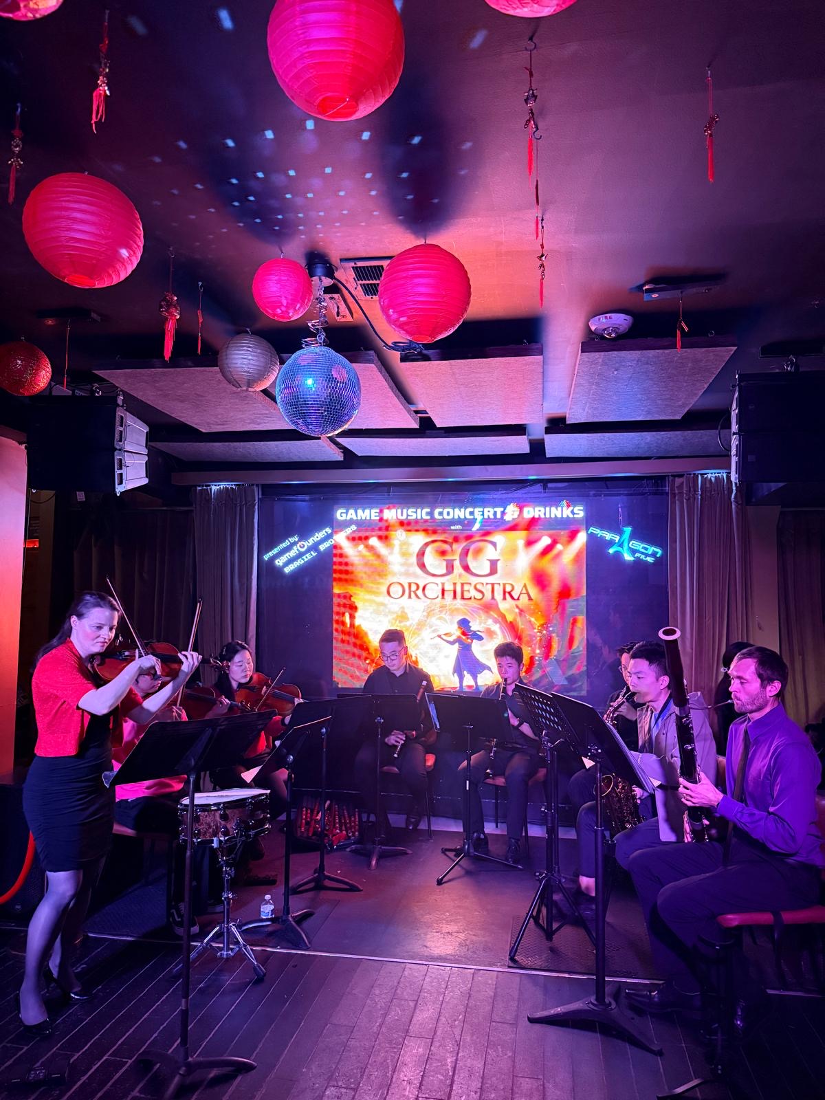 Full ensemble performing at Lion's Den with GG Orchestra banner and atmospheric lighting
